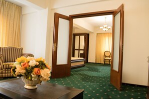 Habitación de hotel con alfombra verde, puerta de madera, cama con cabecera blanca, una silla y un florero con flores sobre una mesa.
