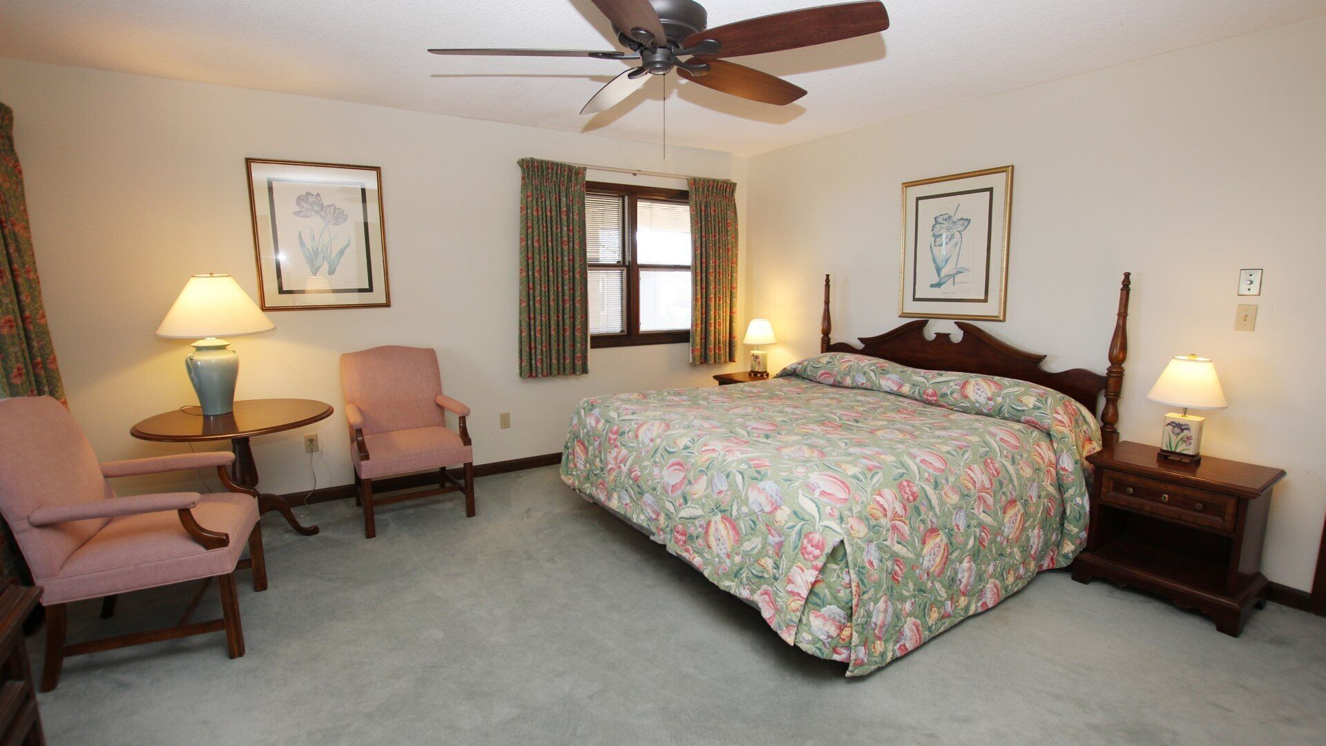 A bedroom with a bed, two chairs, a round table, a ceiling fan, and two lamps.