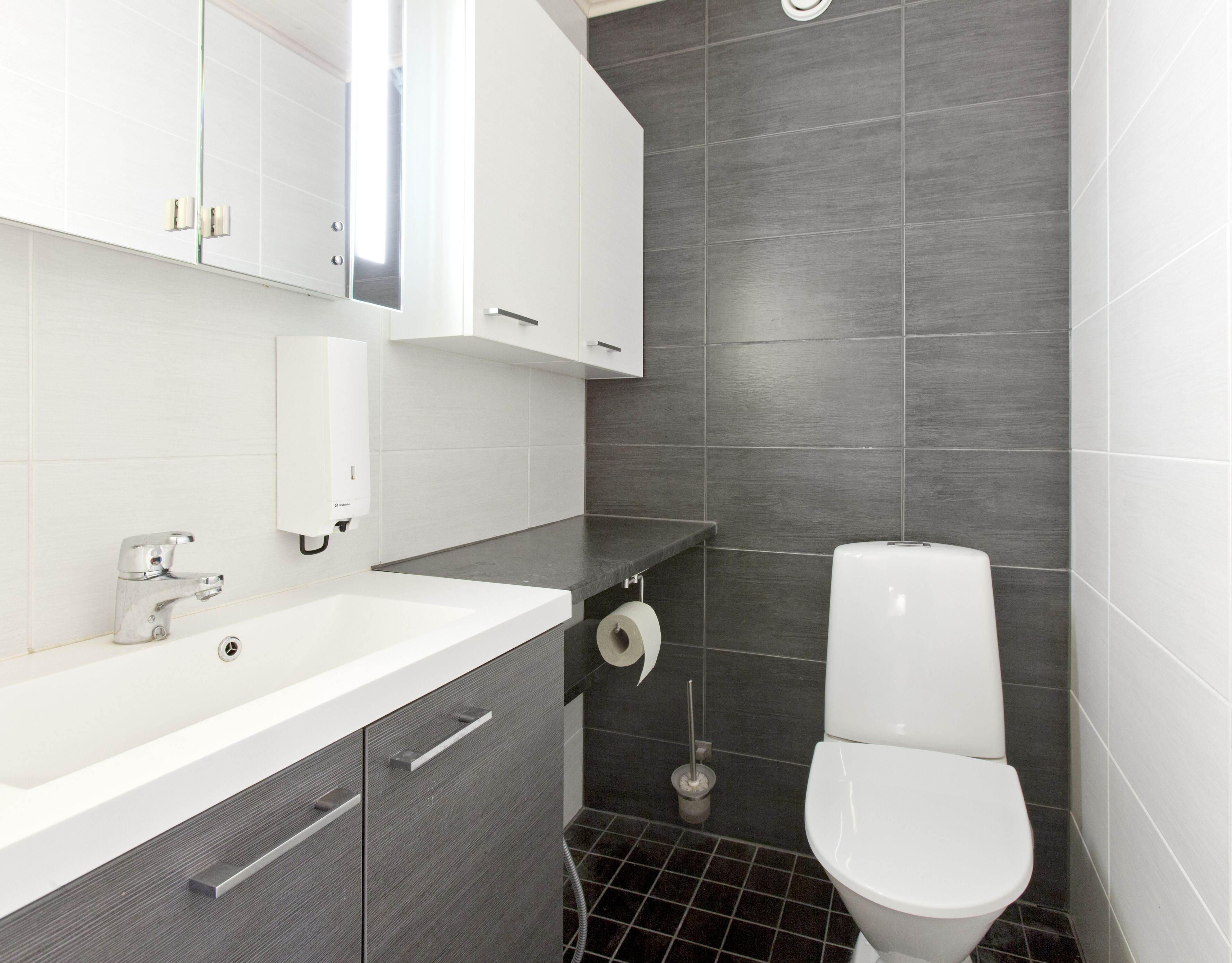 A modern bathroom with a white sink, dark grey tiles, a toilet, and a mirror above the sink.