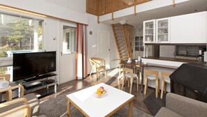 Un salon moderne doté d’un téléviseur à écran plat, d’un coin repas avec des chaises en bois, d’une cuisine avec des meubles blancs et d’un escalier.