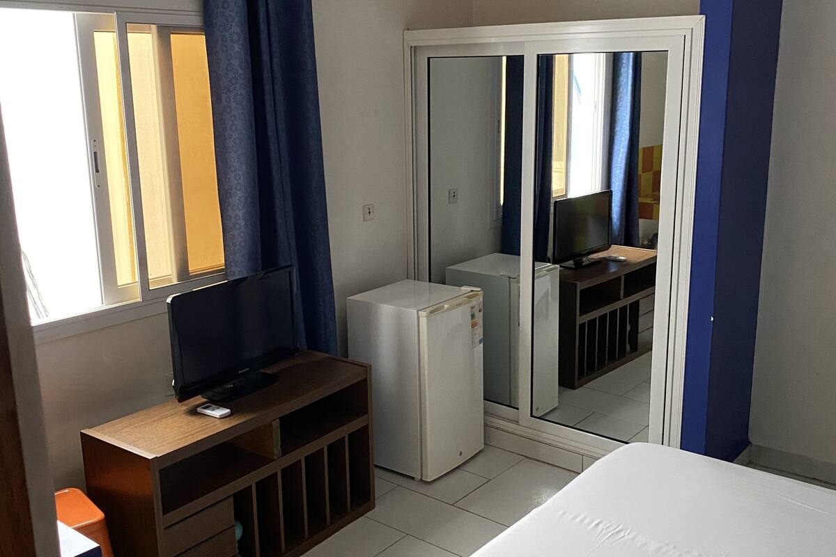A bedroom with a bed, a TV, a small fridge, a mirror, and a window with curtains.