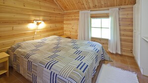Un dormitorio tipo cabaña de madera con una cama, una mesita de noche, un ventanal con cortinas y techo de madera.