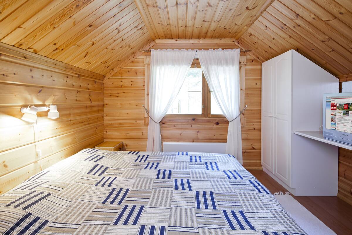 Ein Schlafzimmer einer Holzhütte mit einem Bett, einem Fenster mit Vorhängen, einem weißen Kleiderschrank und einer Wandleuchte.