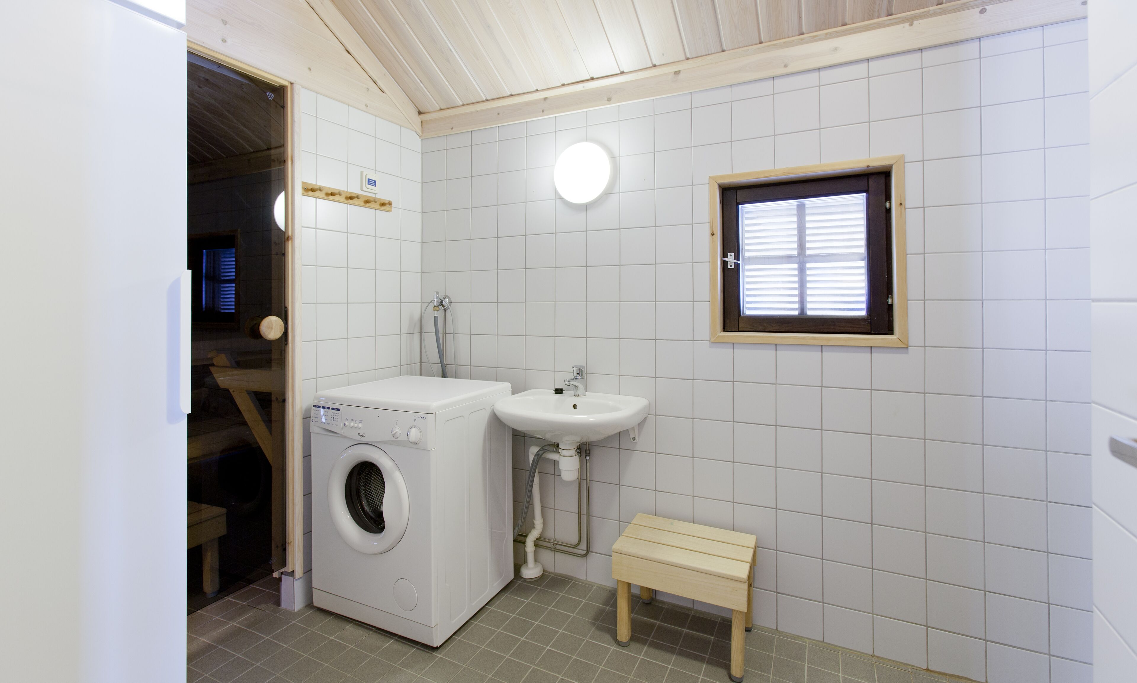 Un baño pequeño y bien iluminado con lavadora, lavamanos y un taburete de madera.