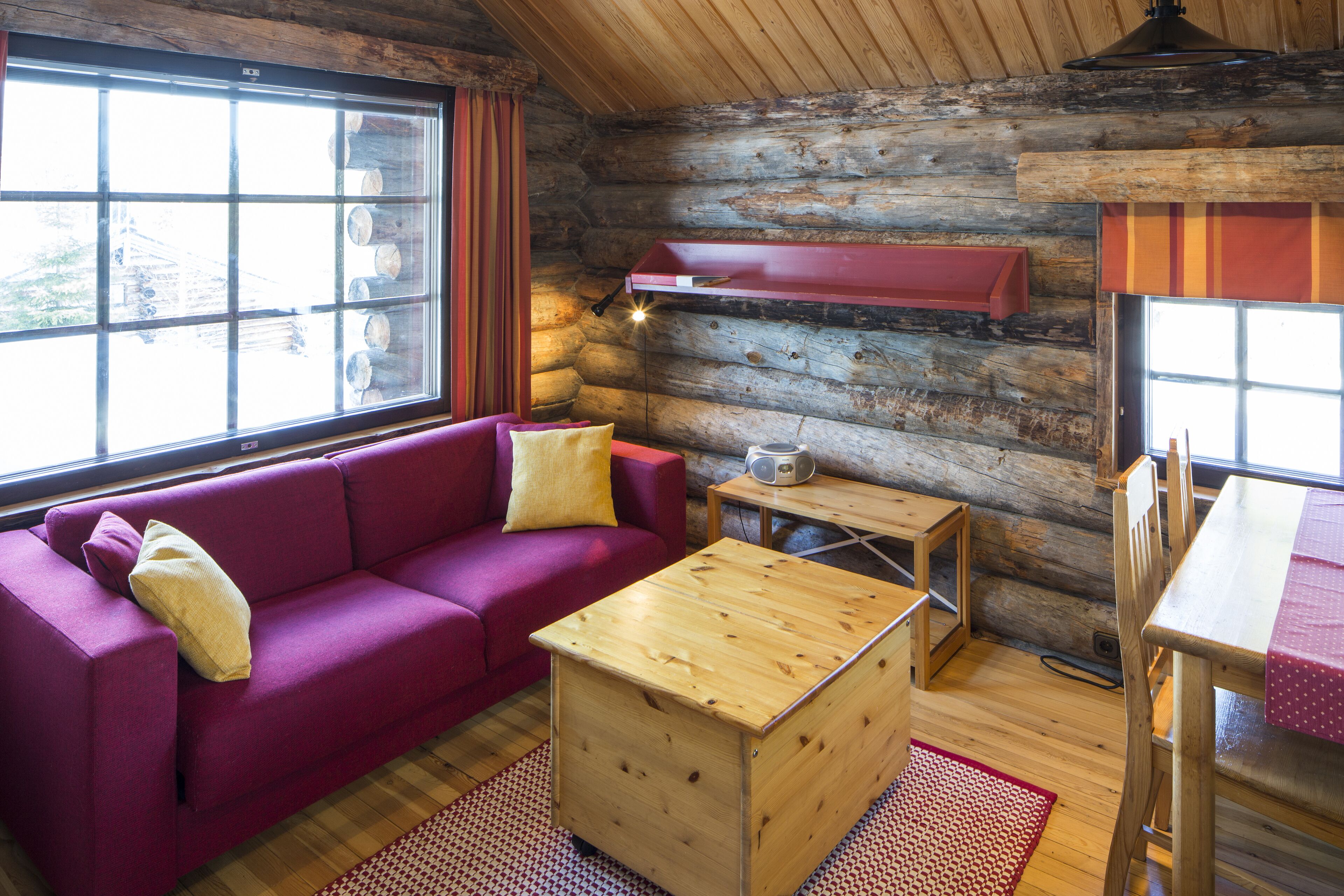 A log cabin interior with a red sofa, wooden coffee table, and a dining table with a red tablecloth.