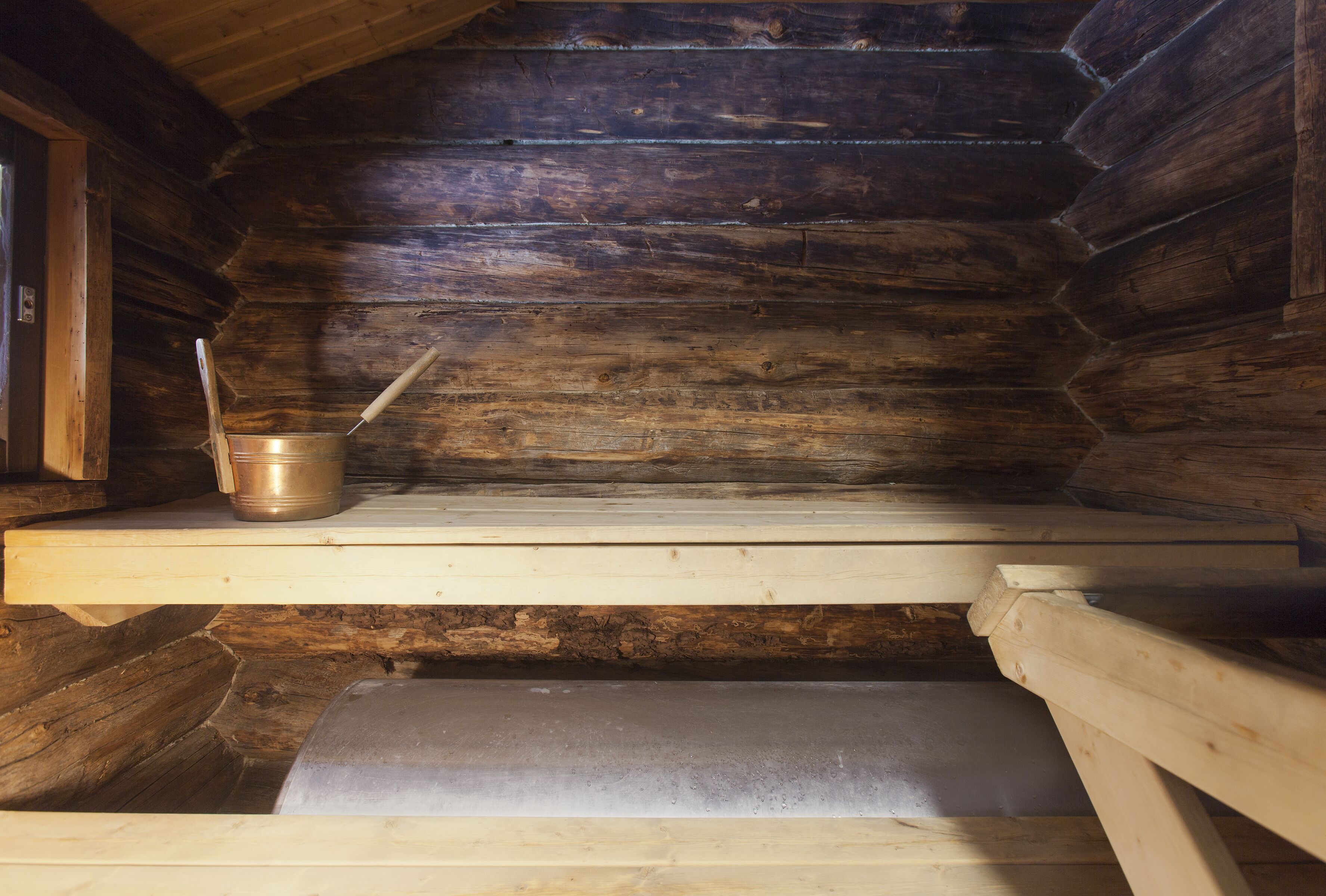 A wooden cabin interior with a metal stove, a wooden bench, and a shelf with a metal pot.