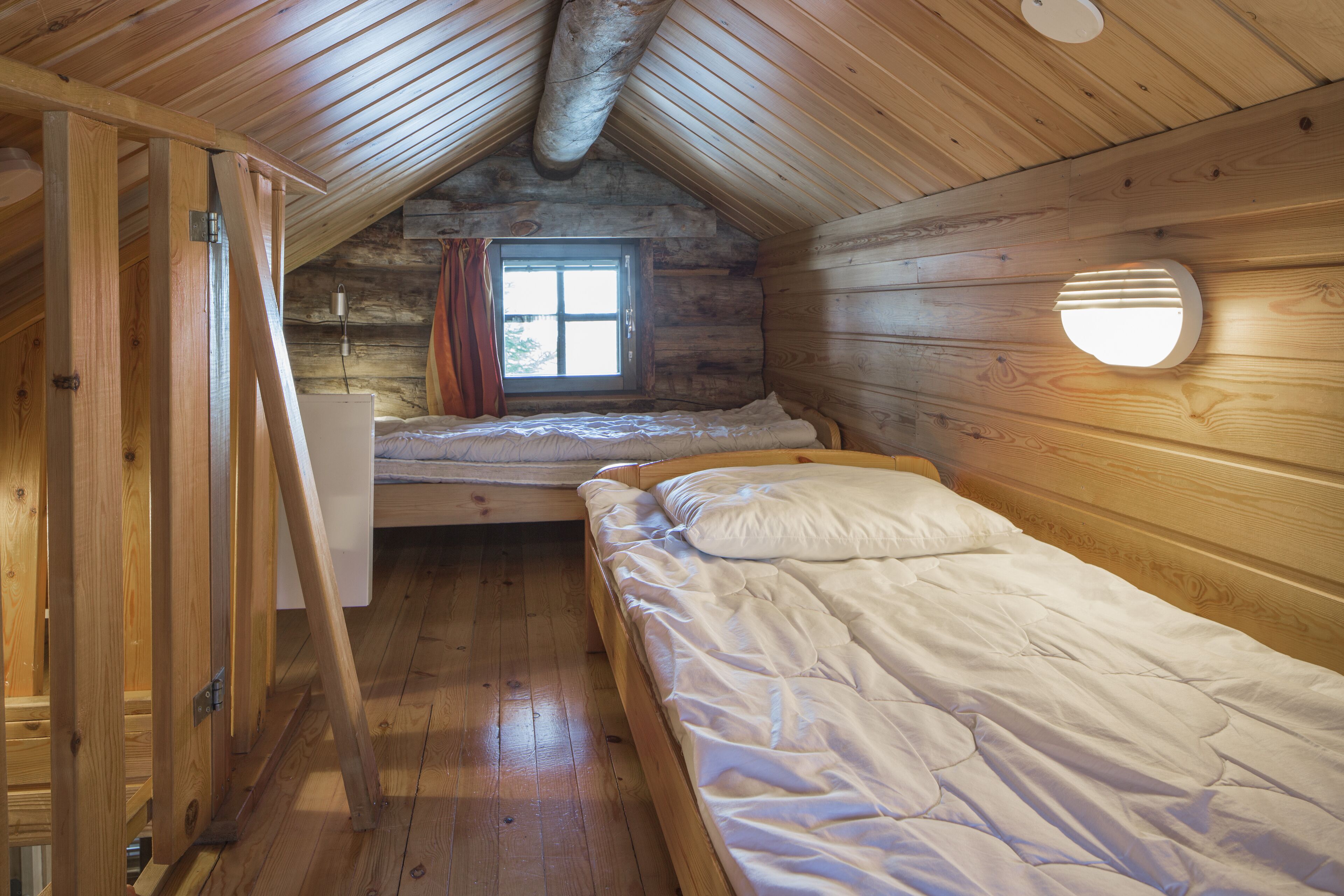 Une petite pièce agréable, avec un plafond en pente, des murs en bois, un lit simple, une fenêtre avec des rideaux et un luminaire fixé au mur.
