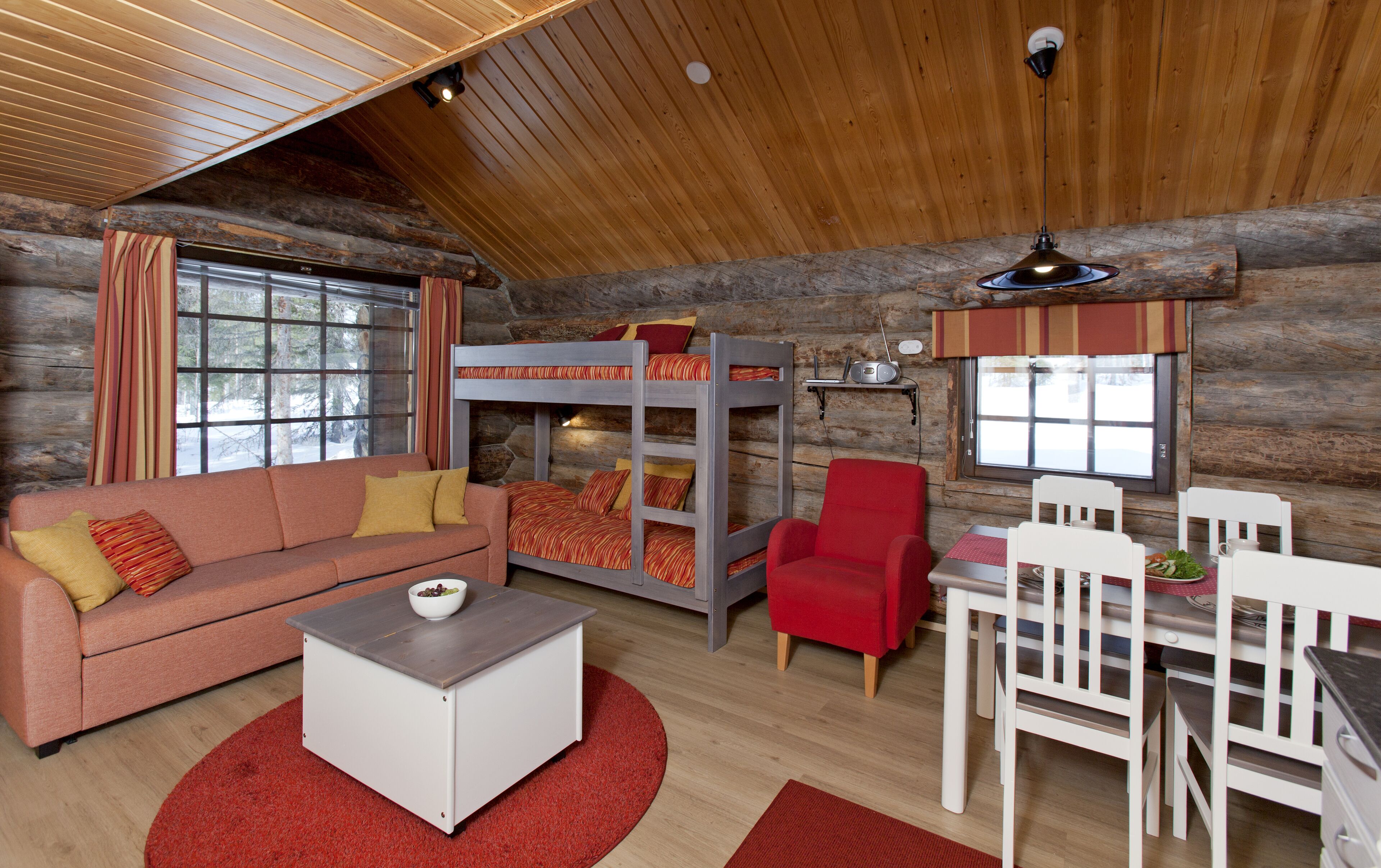 A cozy cabin interior with a bunk bed, a sofa, a dining table, and a red rug.