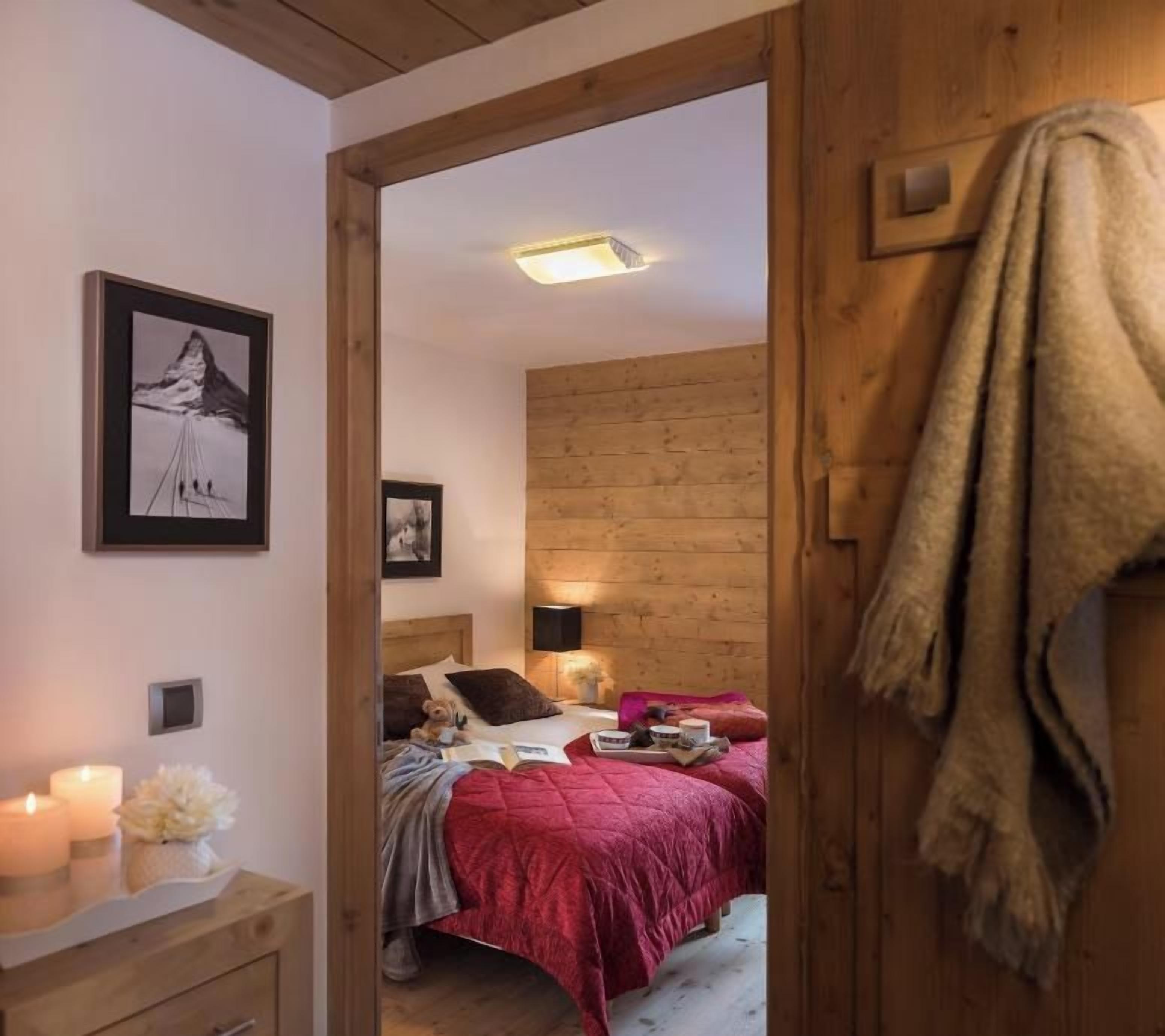 Une chambre avec une porte en bois, un lit recouvert d’une couverture rouge, une table de chevet avec une bougie et un tableau encadré accroché au mur.