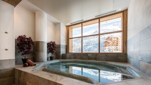 A spa area with a hot tub, potted plants, and a view of a mountain resort through a large window. - CGH Le Lodge des Neiges (Tignes)