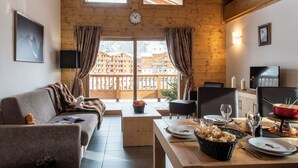 A cozy room with a sofa, dining table, and a view of a mountain resort. - CGH Le Lodge des Neiges (Tignes)
