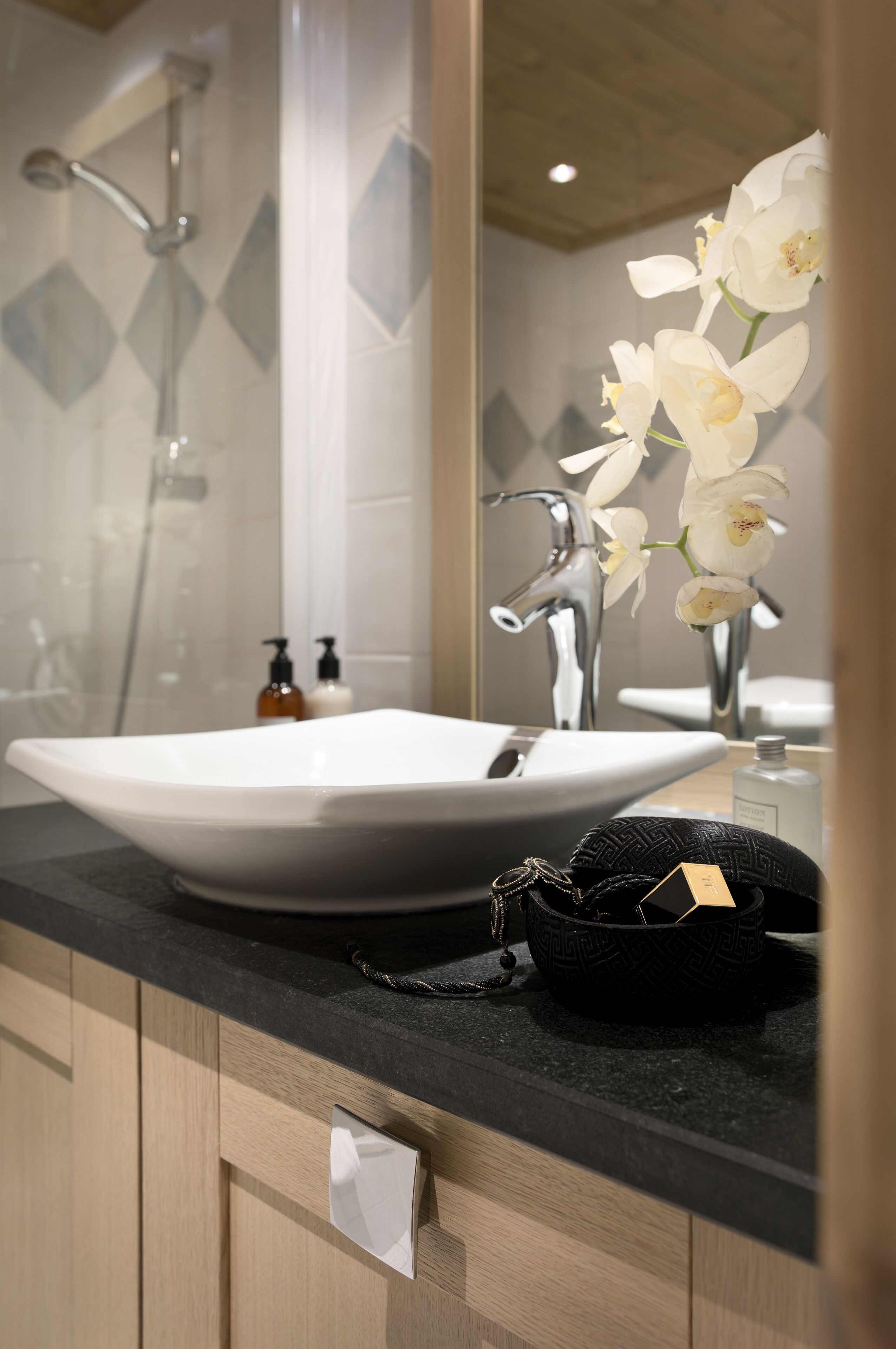 A bathroom with a white sink, a black countertop, and a mirror reflecting a shower and orchid flowers.