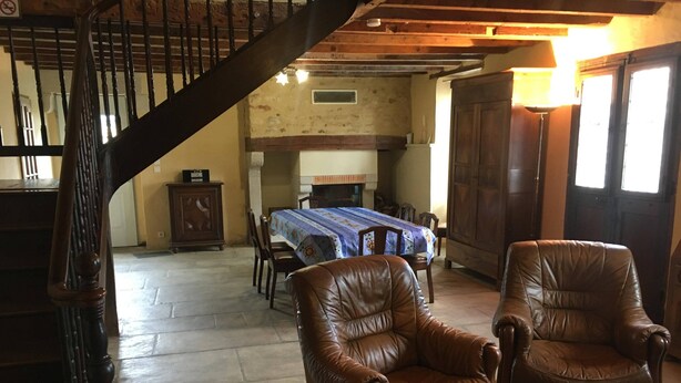 A spacious living room with a high ceiling, a wooden staircase, leather armchairs, a dining table with chairs, and a fireplace.