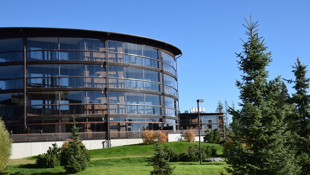Un bâtiment moderne doté d’une façade en verre incurvée, entouré d’un espace paysager agrémenté d’arbres et d’un sentier.