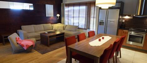 A modern living room with a sofa, coffee table, and dining table with chairs. There is a kitchen area with a stove and oven.