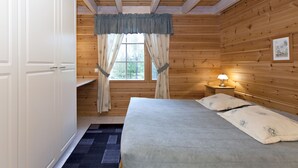 A wooden cabin room with a bed, a nightstand with a lamp, a window with curtains, and a door.