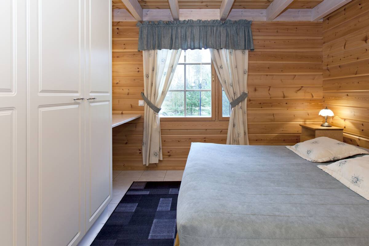 A wooden cabin room with a bed, a nightstand with a lamp, a window with curtains, and a door.