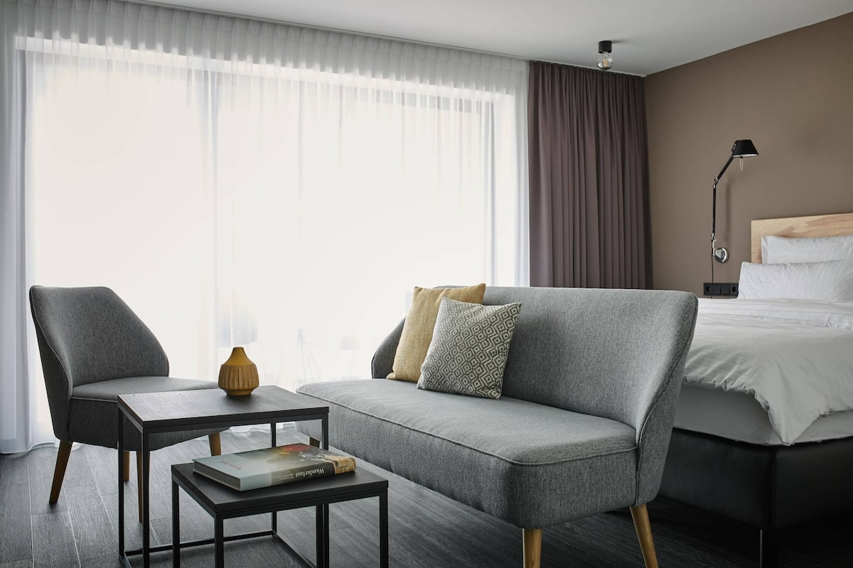 Ein modernes Hotelzimmer mit grauem Sofa, einem kleinen Tisch mit einem Buch, einem Bett mit weißer Bettwäsche und einem Nachttisch mit Lampe.