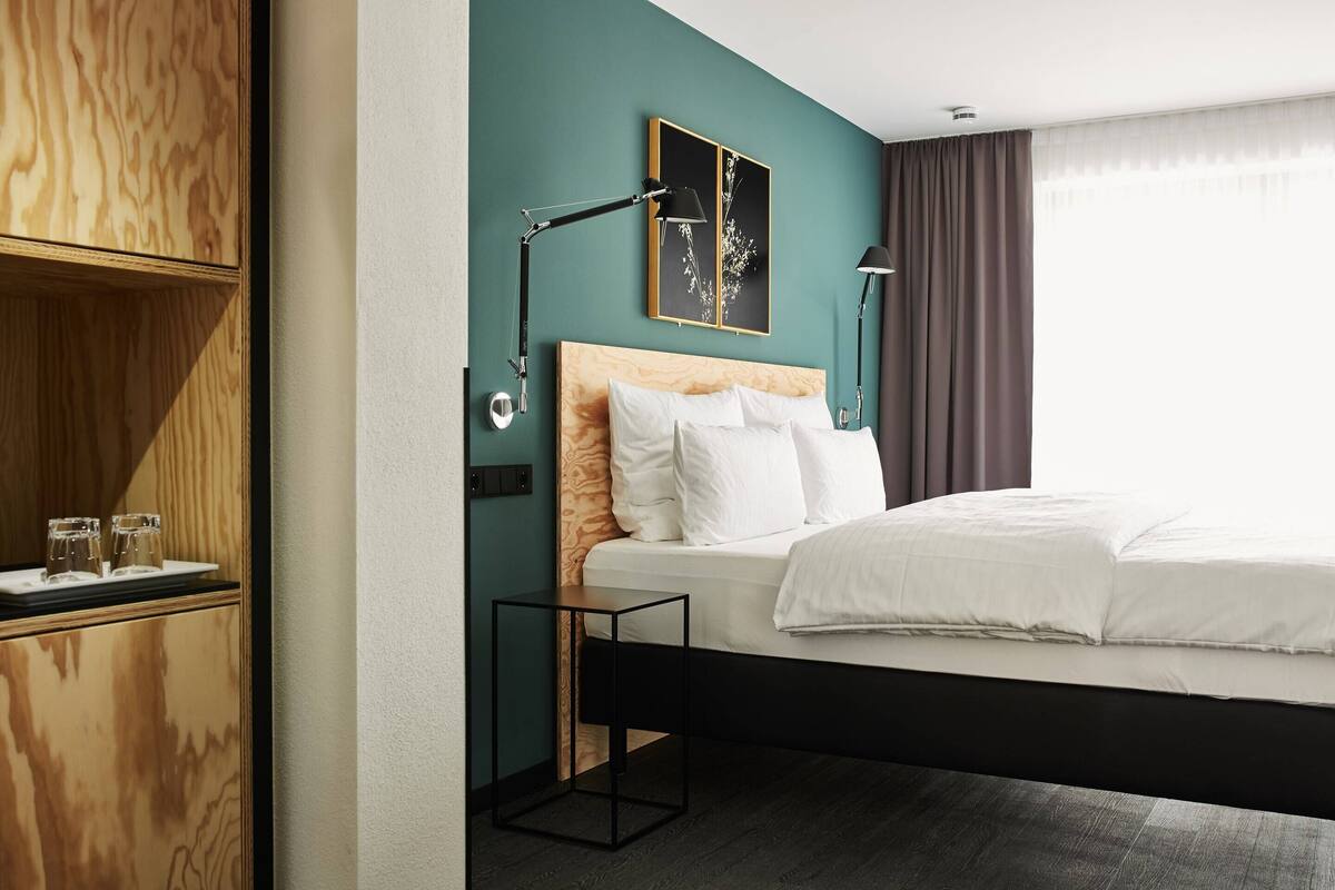 Ein modernes Hotelzimmer mit einem hölzernen Kopfteil, einem Bett mit weißen Laken, einem Nachttisch mit Lampe und einem an der Wand befestigten Bild.