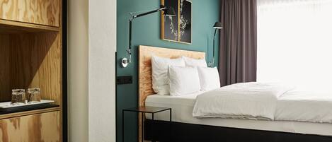 Habitación de hotel moderna con cabecera de madera, cama con ropa blanca, mesita de noche con lámpara y un cuadro colgado en la pared.