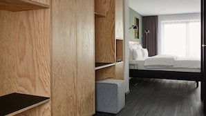 A hotel room with a wooden wardrobe, a grey ottoman, a bed with white linens, and a large window with curtains. - Winterfeld Guest House Bodenmais (Bodenmais)
