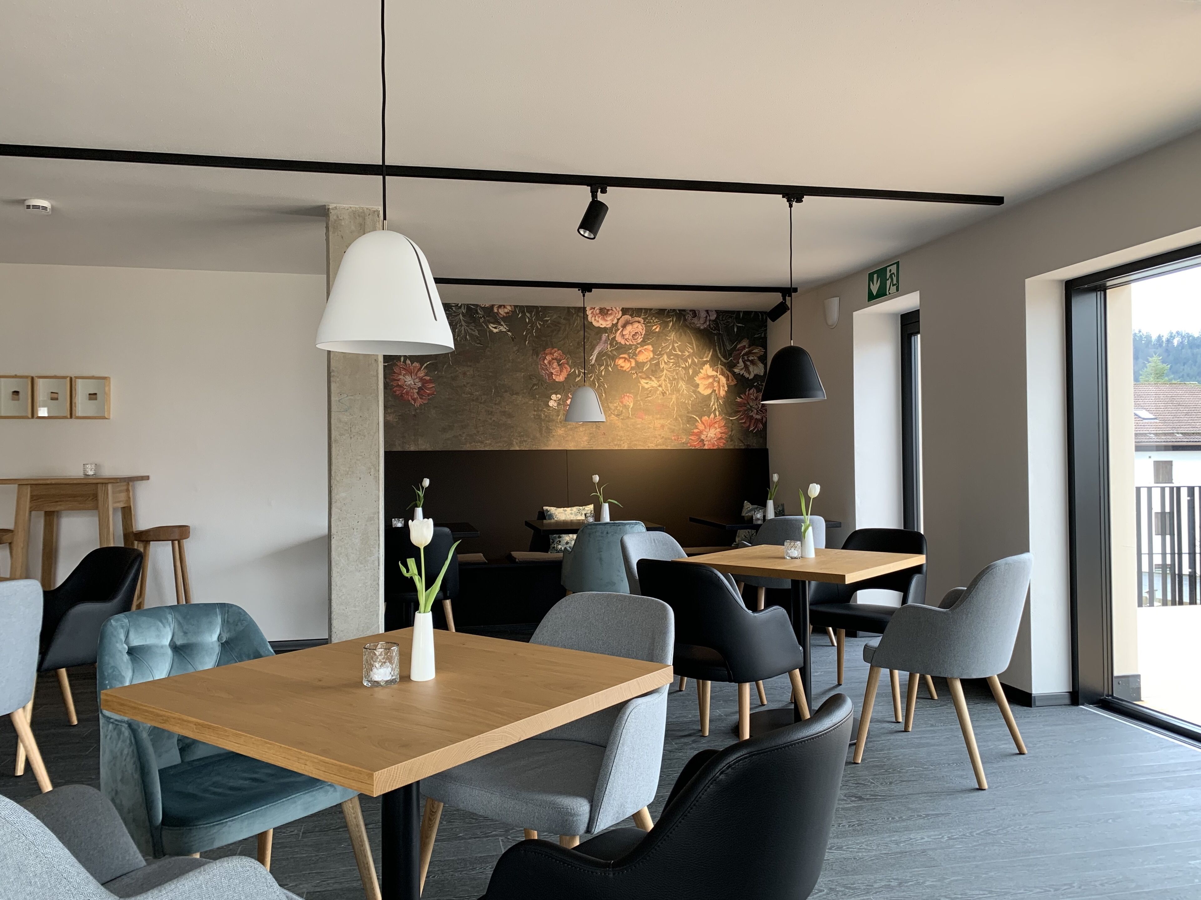 Un comedor moderno con mesas de madera y sillas tapizadas de gris, un mural floral y un ventanal que ofrece vistas al exterior.