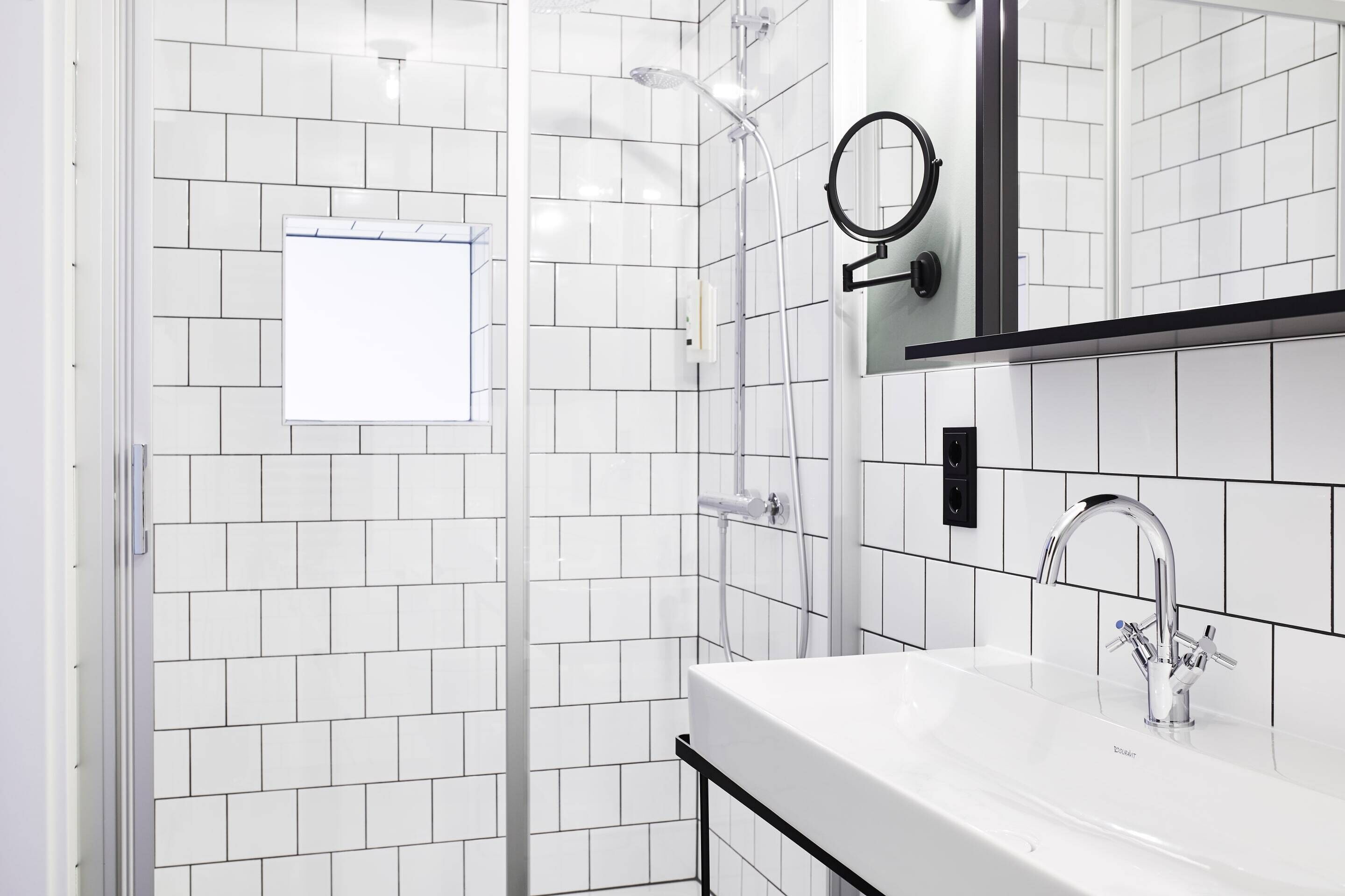 Een moderne badkamer met witte betegelde wanden, een glazen douchecabine, een witte wastafel met chromen kraan en een spiegel met ronde lijst.