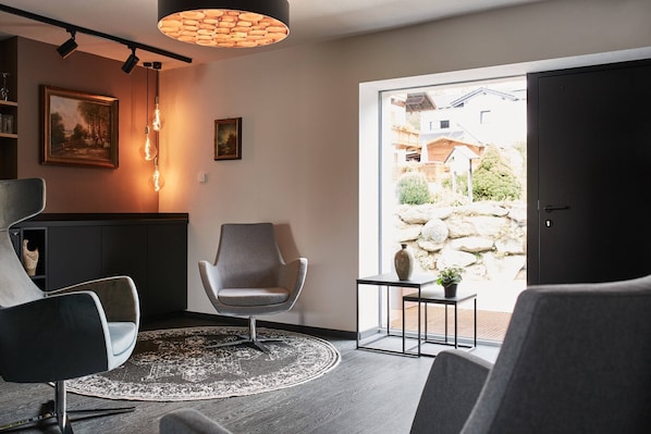A modern living room with a grey armchair, a round rug, and a view of a garden through a sliding door. - Winterfeld Guest House Bodenmais (Bodenmais)