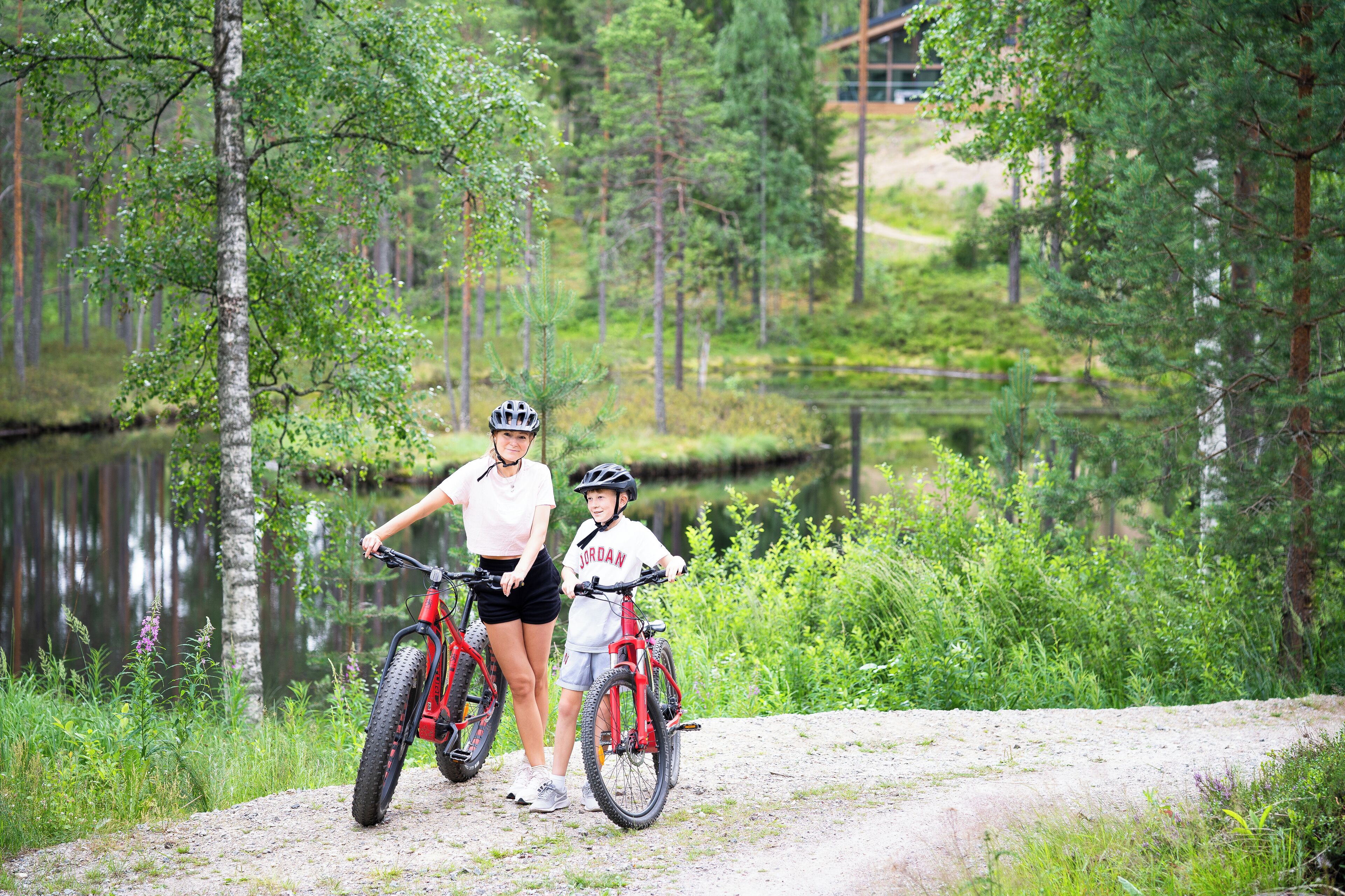Henkilö ja lapsi seisovat vierekkäin polkupyörien kanssa maastopolulla, taustalla näkyy järvi ja metsä.