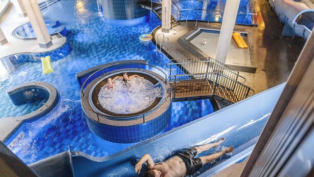 Una persona se desliza por un tobogán acuático en un área de piscina con un jacuzzi y una escalera.