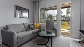 Una sala moderna con un sofá gris, una mesa de centro negra y un sillón gris. Hay un ventanal con vistas a árboles y una terraza de madera.