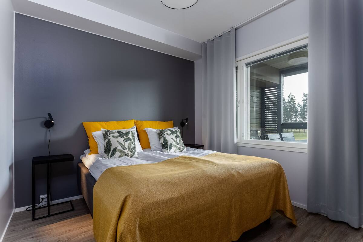 A modern bedroom with a bed, a bedside table, a window with curtains, and a view of trees outside.