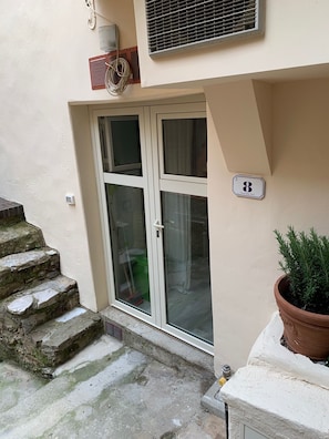 A building entrance with a glass door, a small potted plant, and a number sign indicating the unit.