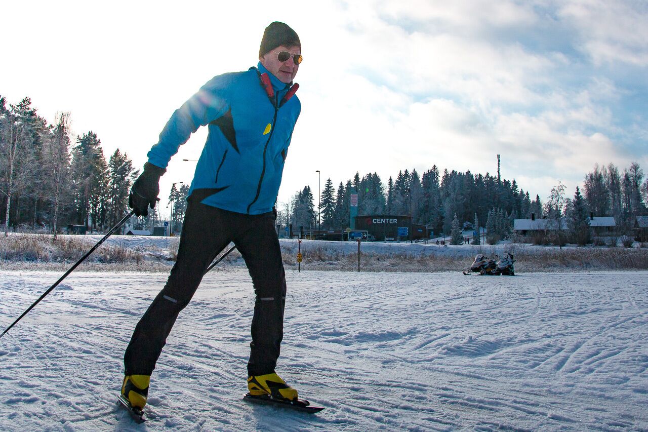 A person cross-country skiing on a snowy field with a sled in the background.