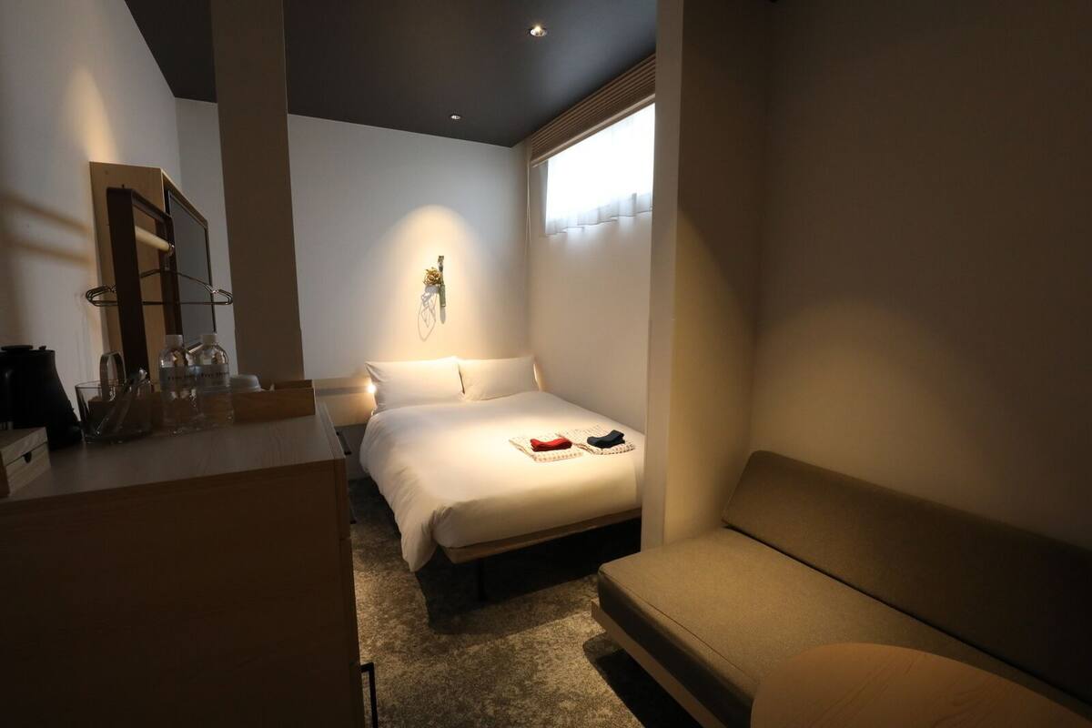 A modern hotel room with a bed, a small desk, a chair, and a window.
