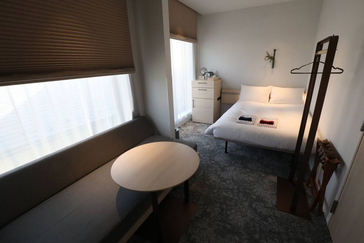 A hotel room with a bed, a bedside table, a dresser, and a window with blinds.