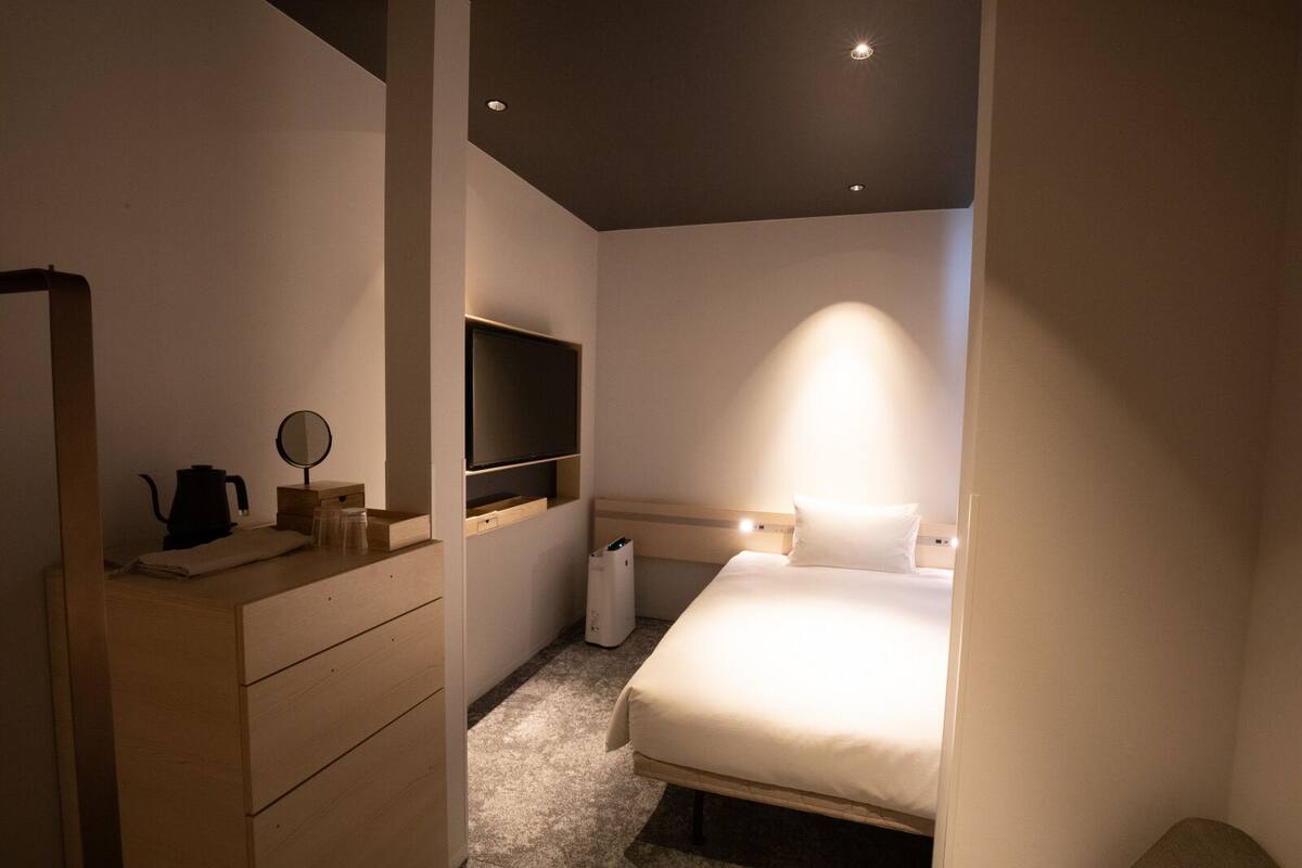 A modern hotel room with a bed, a TV mounted on the wall, a bedside table with a kettle, and a mirror.