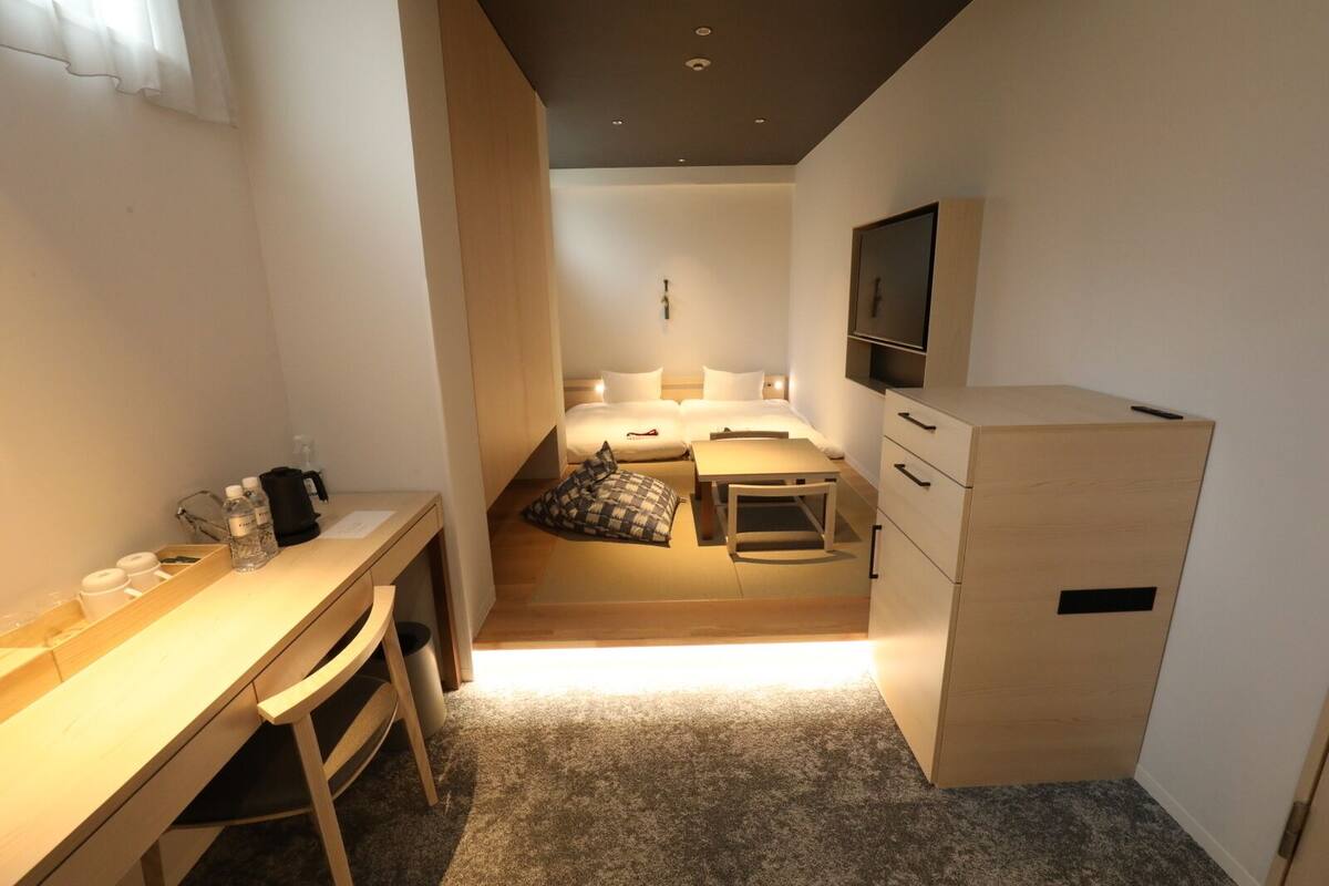 A compact hotel room with a bed, a desk, a chair, a small table, a TV, and a wardrobe.