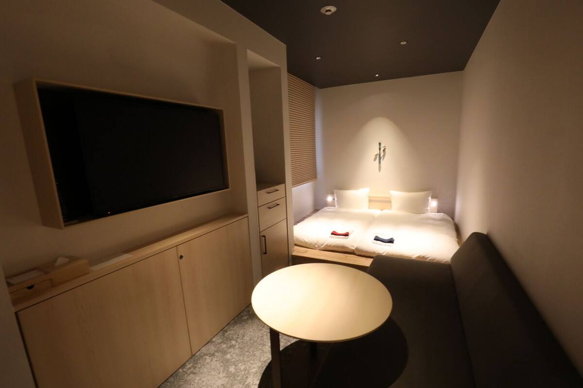 A modern hotel room with a flat-screen TV, a bed with two pillows, a bedside table, and a small desk area.
