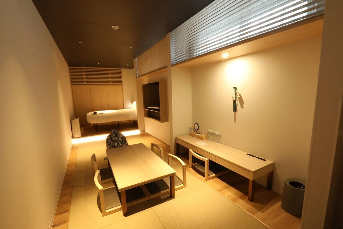 A compact hotel room with a bed, a desk, a chair, a small table, a trash bin, and a wall-mounted shelf with toiletries.