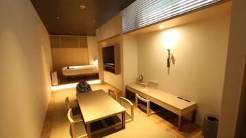 A compact hotel room with a bed, a desk, a chair, a small table, a trash bin, and a wall-mounted shelf with toiletries.