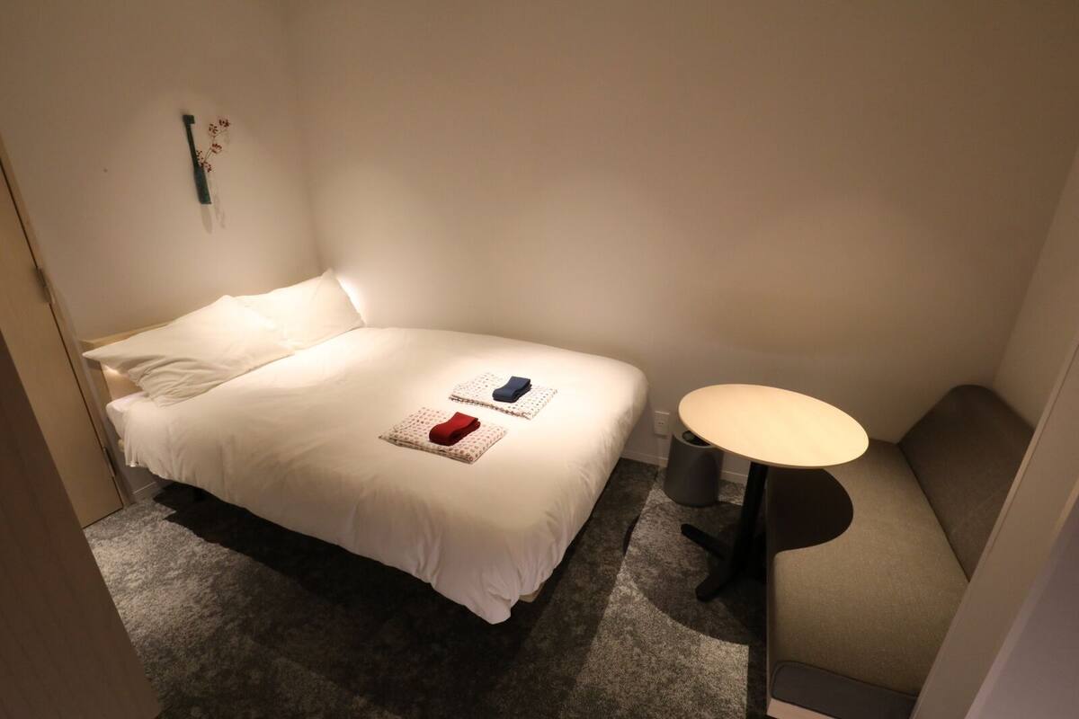 A neatly made bed with a pillow, a small round table, and a wall-mounted light fixture.