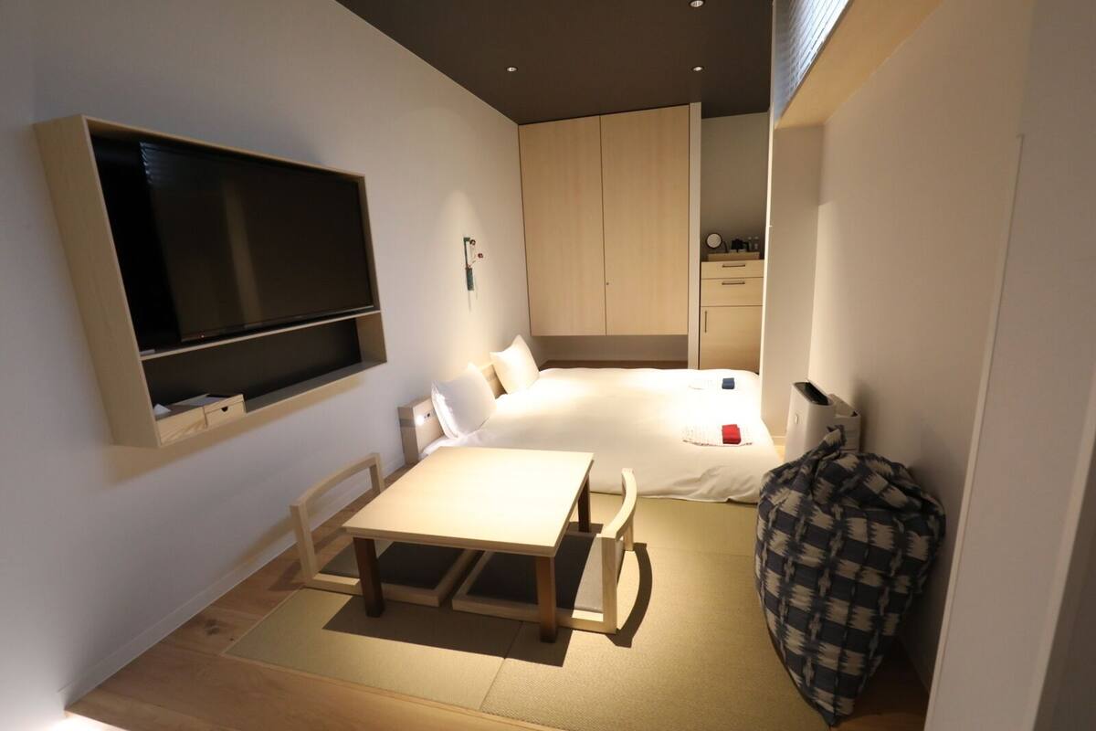 A compact hotel room with a bed, a small table, a TV mounted on the wall, and a kitchenette area.