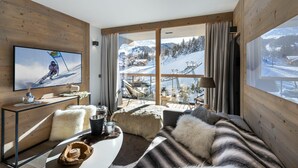 A cozy living room with a large TV, wooden walls, a sofa with fur throw pillows, a coffee table with decorative items, and a view of a snowy mountain landscape through large windows. - Winter Courchevel -- Phoenix 603 (Courchevel)