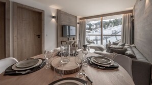 A dining area with a wooden table set for four, a television mounted on the wall, and a view of a snowy landscape through large windows. - Winter Courchevel -- Phoenix 603 (Courchevel)