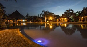 A tranquil pool area at dusk with reflective water, thatched-roof structures, and ambient lighting. - Sandy's Creations Resort (Lusaka)