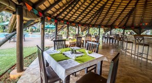 An outdoor dining area with tables set for two, covered by a wooden roof, and surrounded by chairs. - Sandy's Creations Resort (Lusaka)