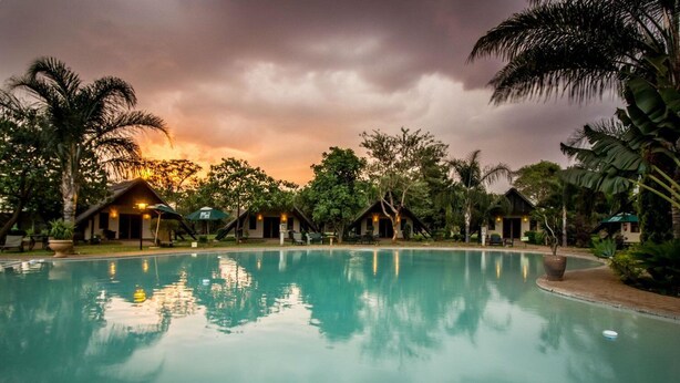 A serene pool area with a sunset view, surrounded by palm trees and small huts. - Sandy's Creations Resort (Lusaka)