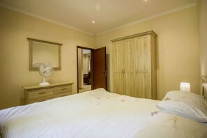 A bedroom with a bed, a wooden wardrobe, a mirror, a bedside table with a lamp, and a fan. - Sandy's Creations Resort (Lusaka)
