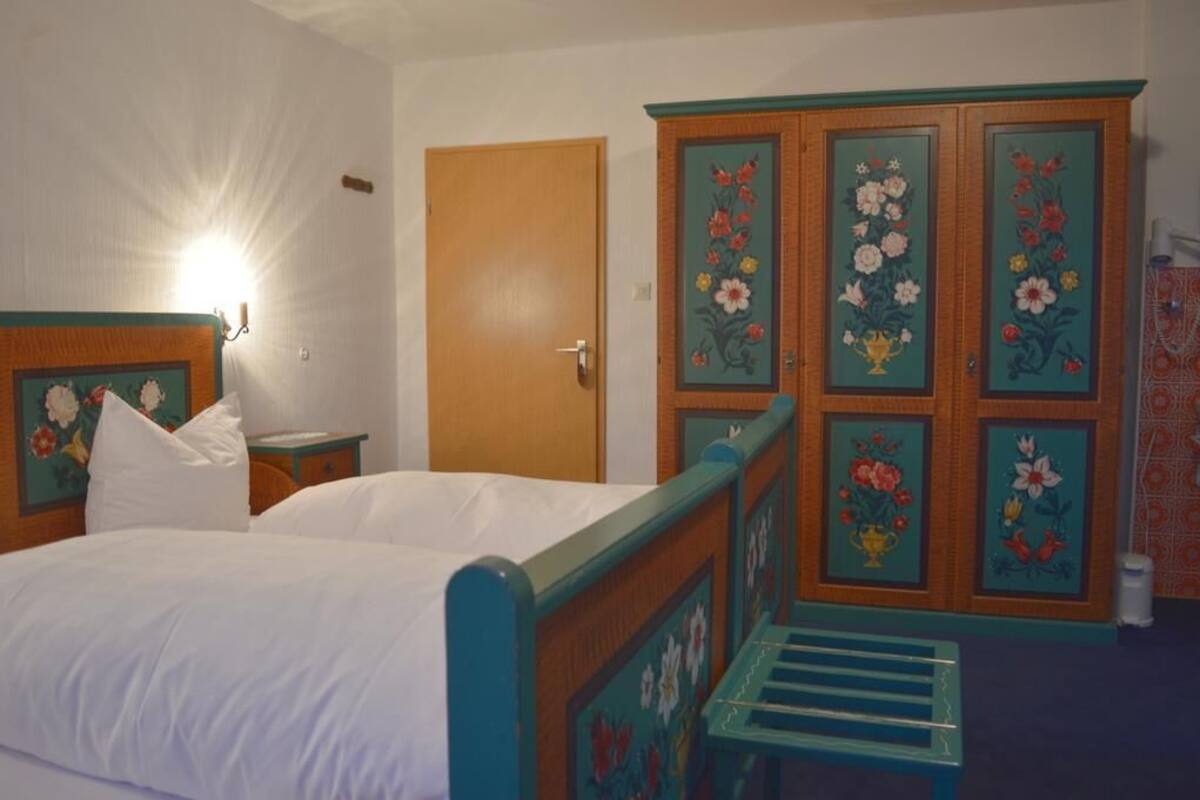 Een slaapkamer met een bed, een nachtkastje, een kledingkast met bloemenmotieven, en een deur.